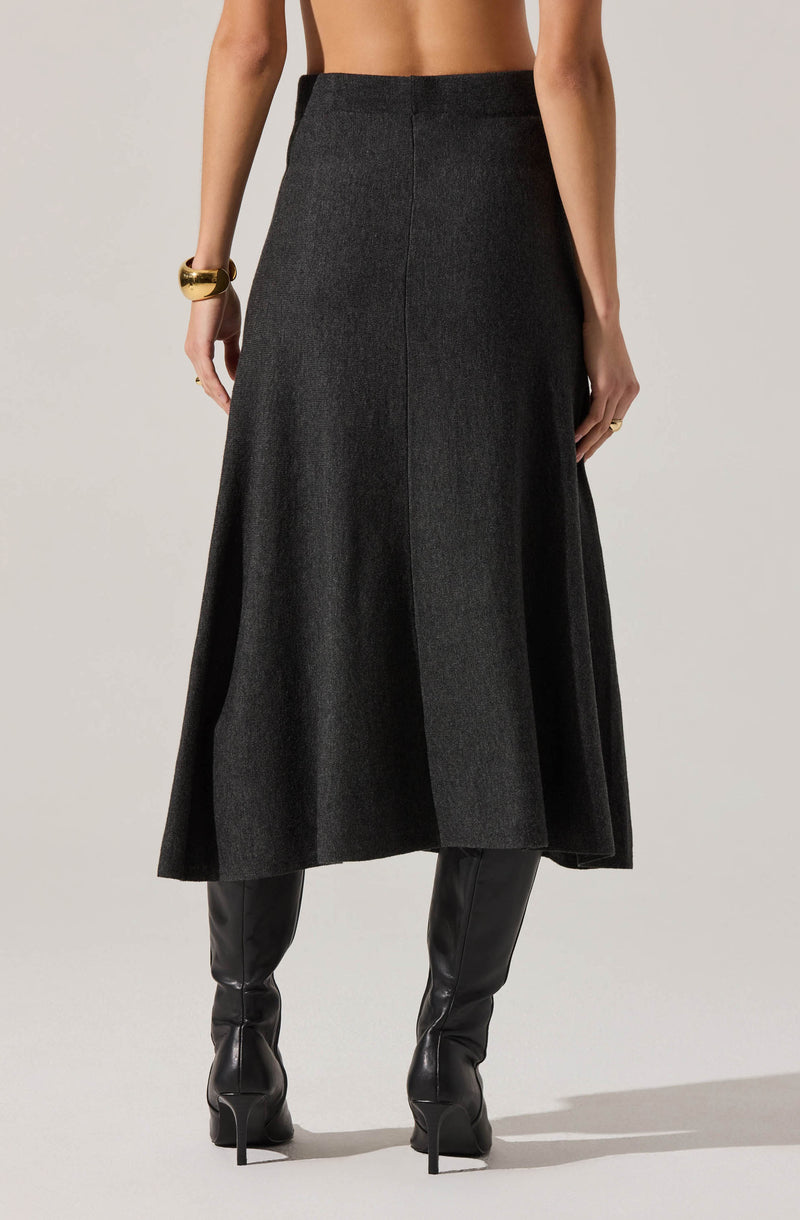 A person is wearing a high-waisted, long, dark gray skirt with a subtle texture. The skirt has a simple, clean design with a front seam running vertically down the center. They are also wearing black, knee-high leather boots with pointed toes and high heels. The person has accessorized with a chunky gold bracelet on their left wrist and a gold ring on their right hand. The background is plain and light-colored.