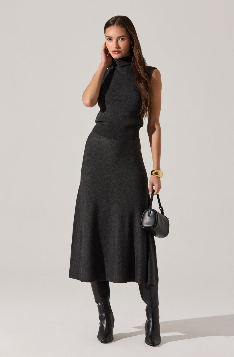 A woman is wearing a sleeveless, high-neck, dark gray knit top paired with a matching long, flowing skirt. She accessorizes with a wide gold cuff bracelet and holds a small black handbag with a short handle. She is also wearing black knee-high boots.