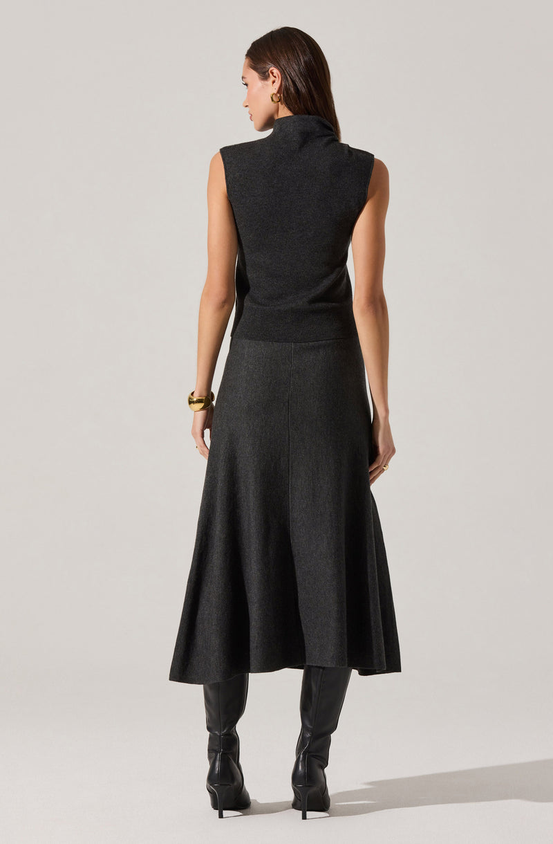 A woman is wearing a sleeveless, high-neck, dark gray knit top paired with a long, flowy, dark gray skirt. She has gold earrings, a gold bracelet on her right wrist, and black high-heeled boots.