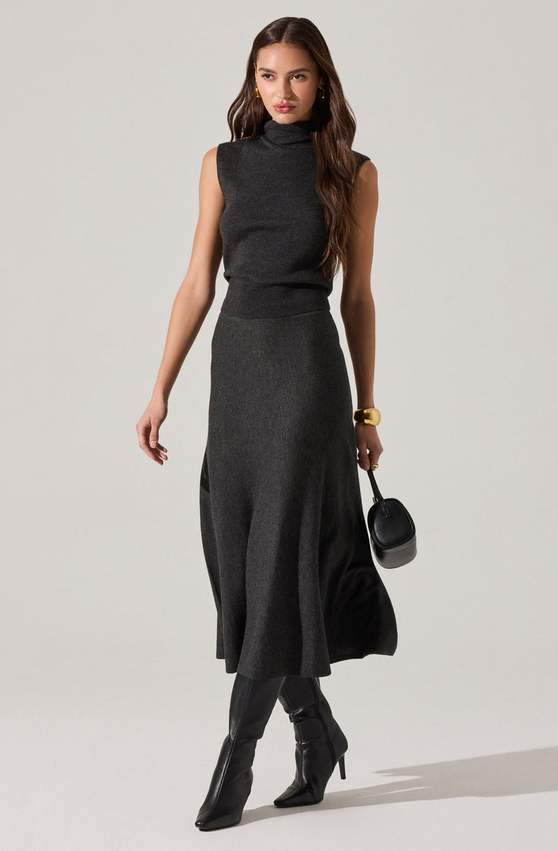A woman is wearing a sleeveless, dark gray turtleneck top and a matching long, flowy skirt in a similar dark gray shade. She accessorizes with a large gold bracelet, gold hoop earrings, and carries a small black handbag. She is also wearing black knee-high boots with high heels.
