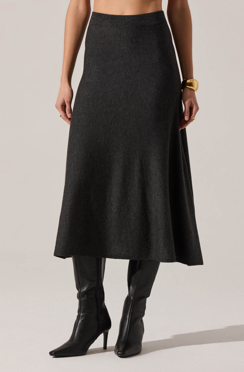 A woman is wearing a long, dark gray skirt with a smooth texture and a high waist. She has paired the skirt with black, knee-high, pointed-toe leather boots with high stiletto heels. She is accessorizing with a large gold bracelet on her right wrist and a ring on her left hand.