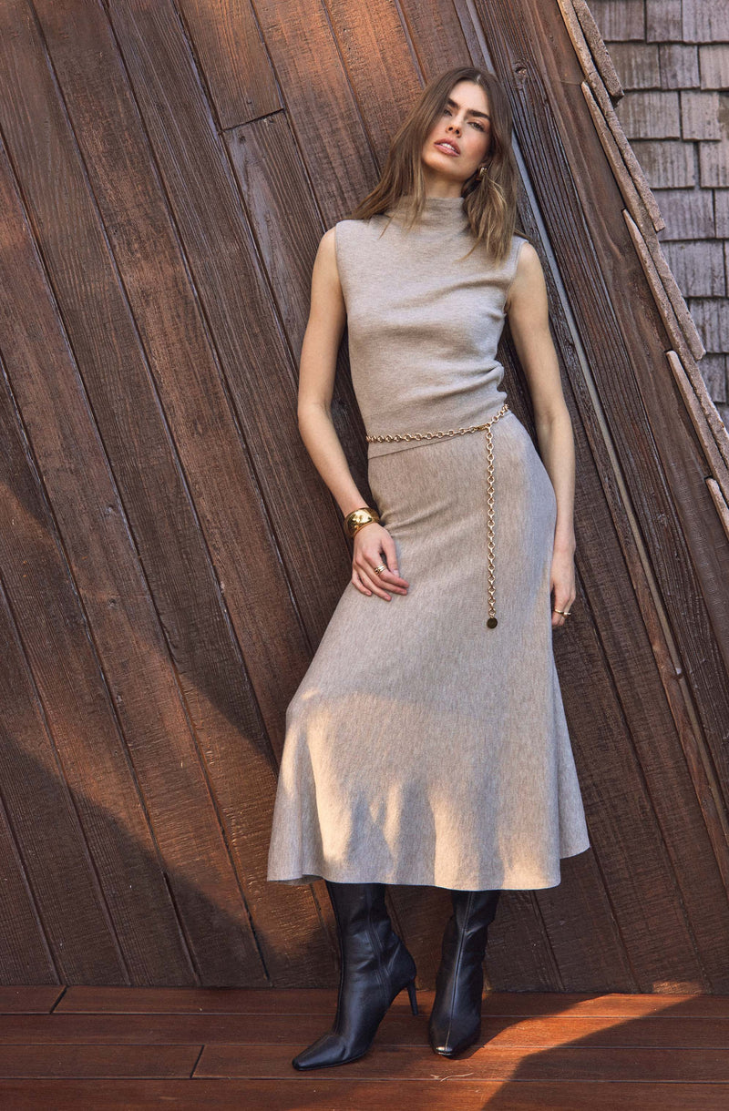 A woman is wearing a sleeveless knit dress in a light beige or oatmeal color that reaches her mid-calf. She accessorized with a gold chain belt hanging loosely around her waist, with one end dangling lower. She has on black high-heeled boots that reach just below the knee. She is also wearing gold bracelets on her left wrist and earrings. Her hair is styled long and loose, and she has natural makeup.
