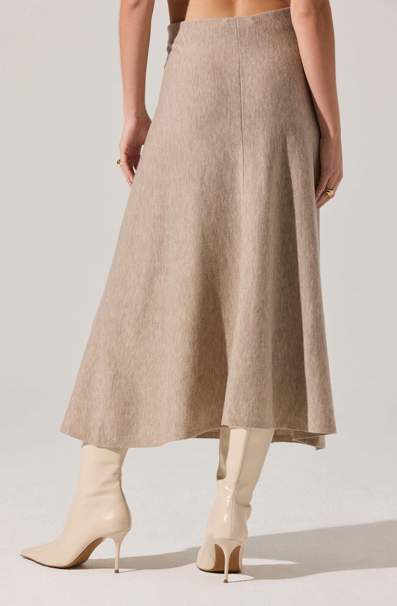 A woman is wearing a high-waisted, ankle-length beige skirt with a subtle texture and a front center seam. She pairs it with white, knee-high, glossy stiletto boots. The image captures her from the waist down, with no top or additional accessories visible.