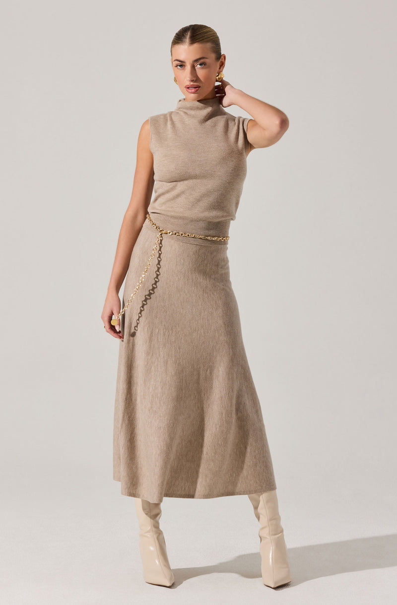 A woman is wearing a sleeveless, high-neck beige knit top paired with a matching beige maxi skirt. She accessorizes with a gold chain belt, gold earrings, and a ring, and is holding a small clutch purse. She is also wearing beige knee-high boots.