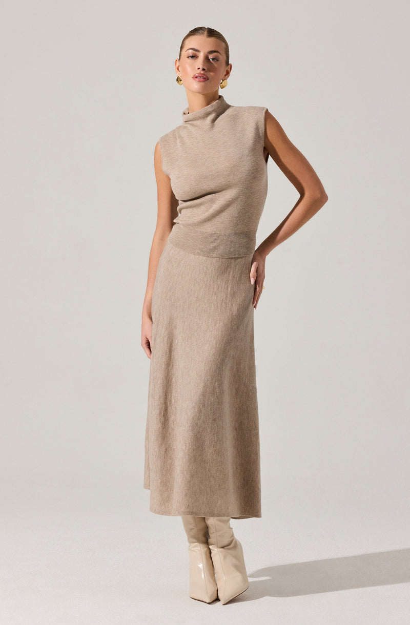 A woman is wearing a sleeveless, high-neck beige knit top with a fitted waist, paired with a long, matching beige knit skirt that reaches her ankles. She is also wearing beige high-heeled boots.