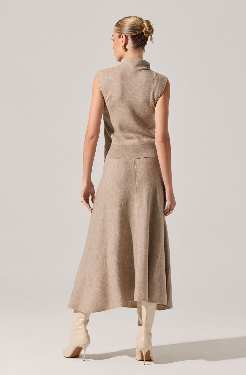 A woman is wearing a beige sleeveless knit vest with a turtleneck collar, paired with a high-waisted, long beige skirt that reaches her ankles. She also has on knee-high beige boots with pointed toes and high heels. Her hair is styled in a neat, low bun.