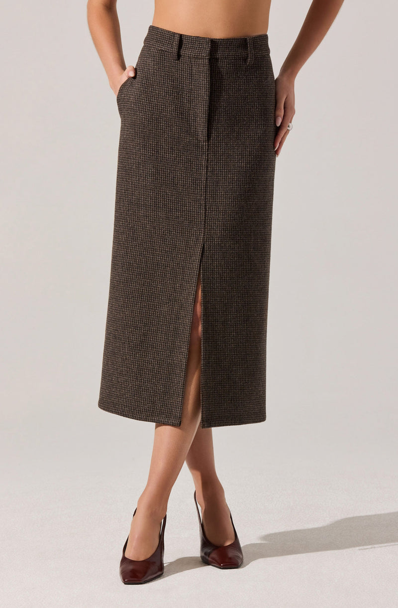 A woman is wearing a high-waisted, ankle-length skirt with a small checkered pattern in brown tones. The skirt features a front slit that extends from the hem to just above the knee. She has her hands in her pockets and is wearing dark burgundy high heels.