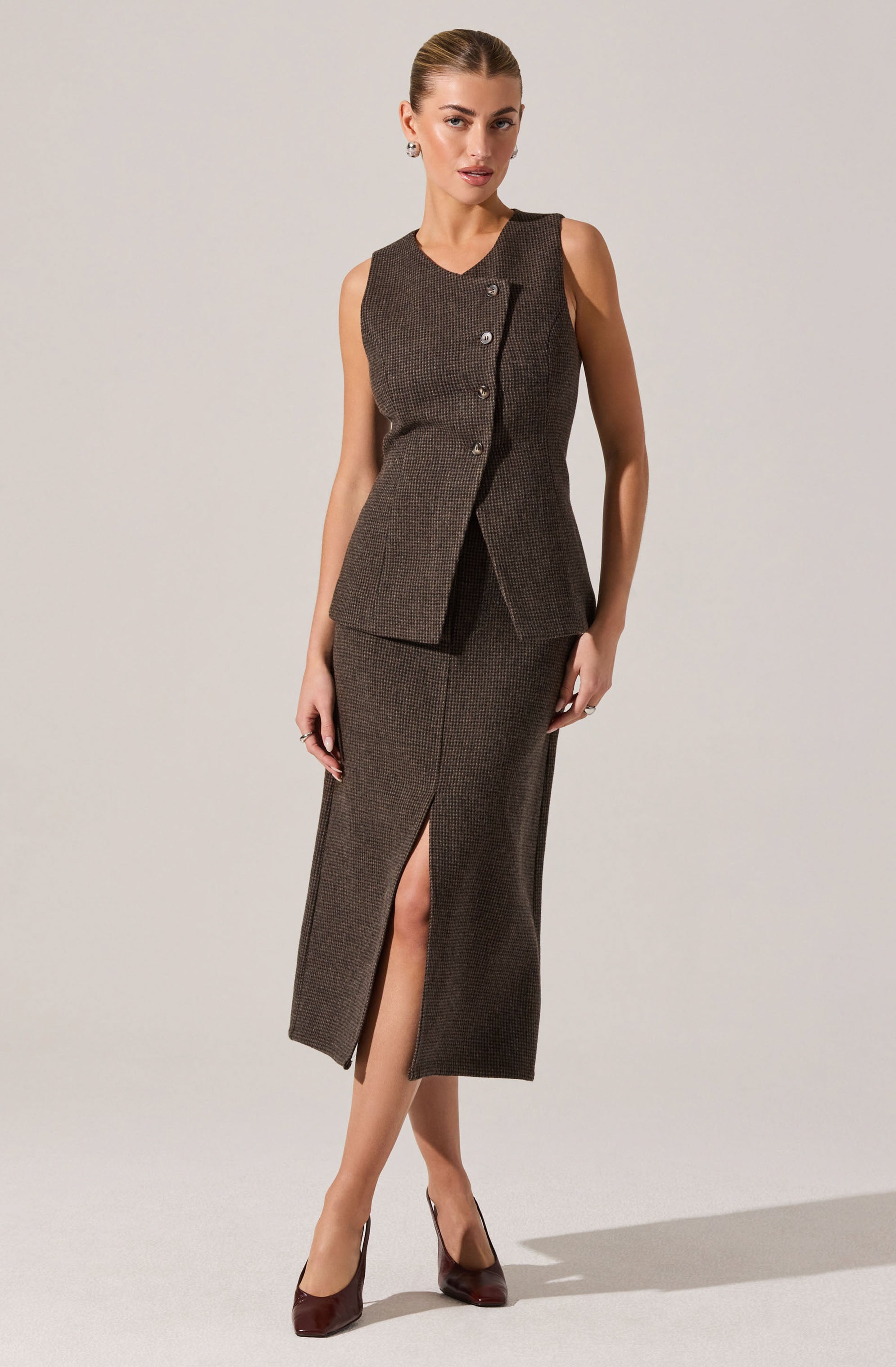 A woman is wearing a sleeveless, fitted brown vest with a subtle checkered pattern, featuring a small collar and three buttons on the front. She pairs it with a matching brown checkered skirt that reaches mid-calf and has a front slit. She completes the outfit with dark brown high-heeled shoes.