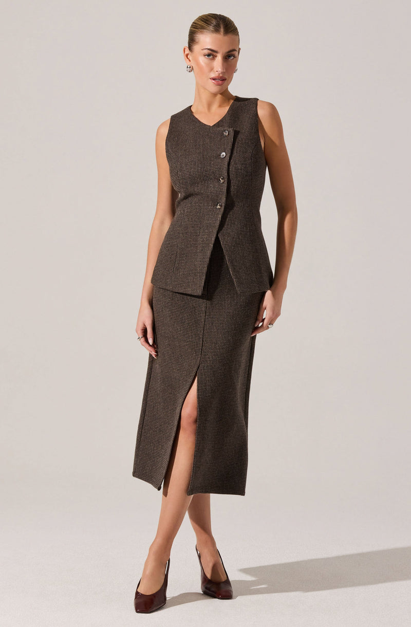 A woman is wearing a sleeveless, fitted brown vest with a subtle checkered pattern, featuring a small collar and three buttons on the front. She pairs it with a matching brown checkered skirt that reaches mid-calf and has a front slit. She completes the outfit with dark brown high-heeled shoes.