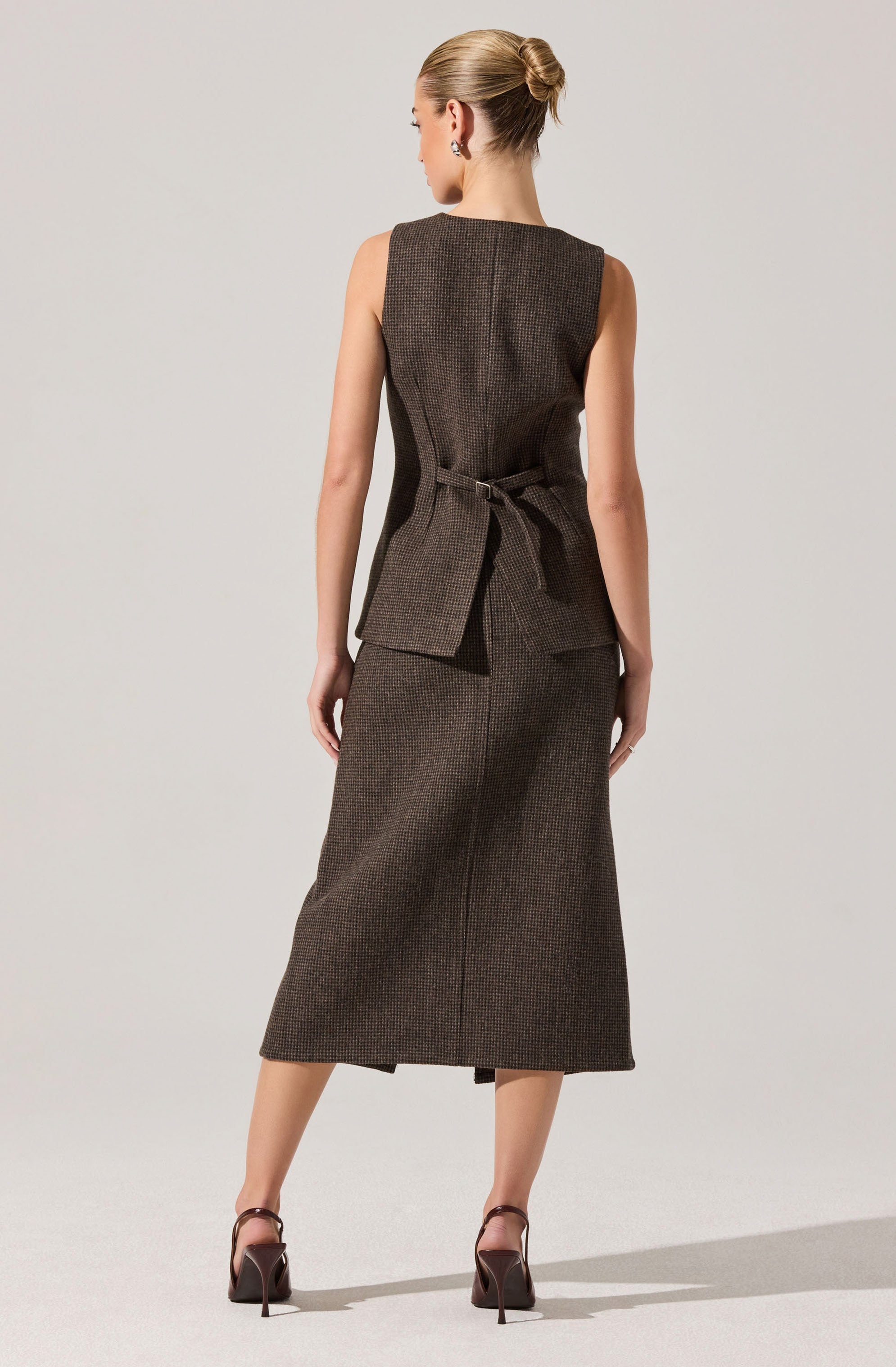A woman is wearing a sleeveless, knee-length, dark brown dress with a subtle checkered pattern. The dress features a tie belt at the waist, creating a fitted silhouette, and has a concealed zipper back closure. She is also wearing high-heeled shoes with ankle straps.