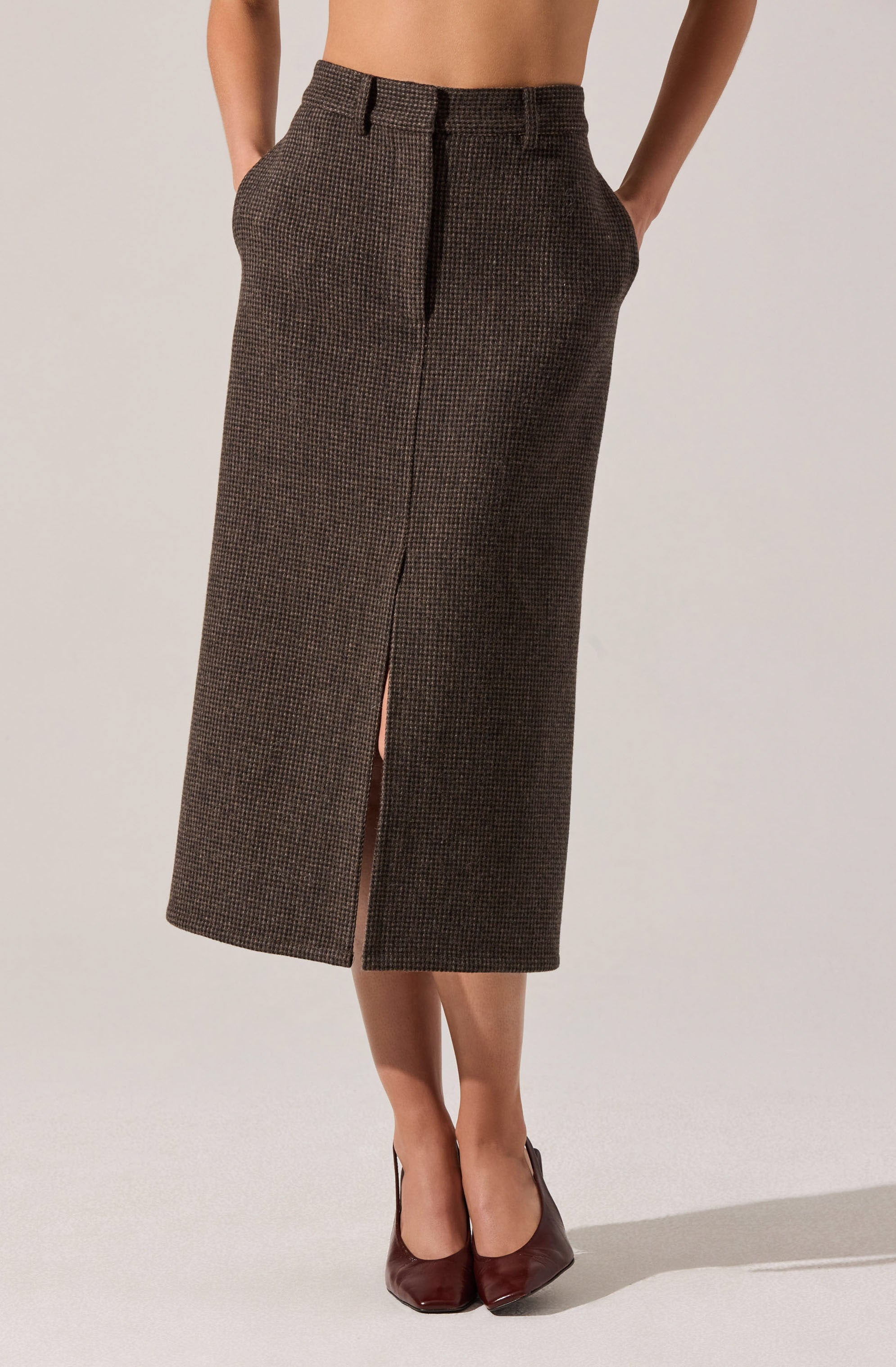 A woman is wearing a pair of high-waisted, straight-cut, knee-length skirt with a small checkered pattern in brown and black. The skirt features a front slit at the center and has a tailored fit. She is also wearing dark-colored high heels.