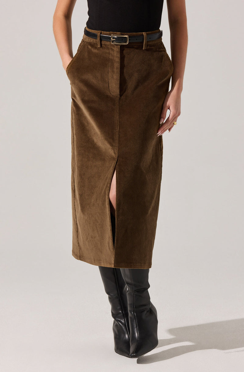 A woman is wearing a black sleeveless top tucked into a high-waisted brown corduroy skirt with a front slit. The skirt has a subtle color variation with a darker segment on one side. She is accessorizing with a black belt that has a rectangular buckle and a gold ring on her right hand. She is also wearing tall black leather boots that reach just below the knee.