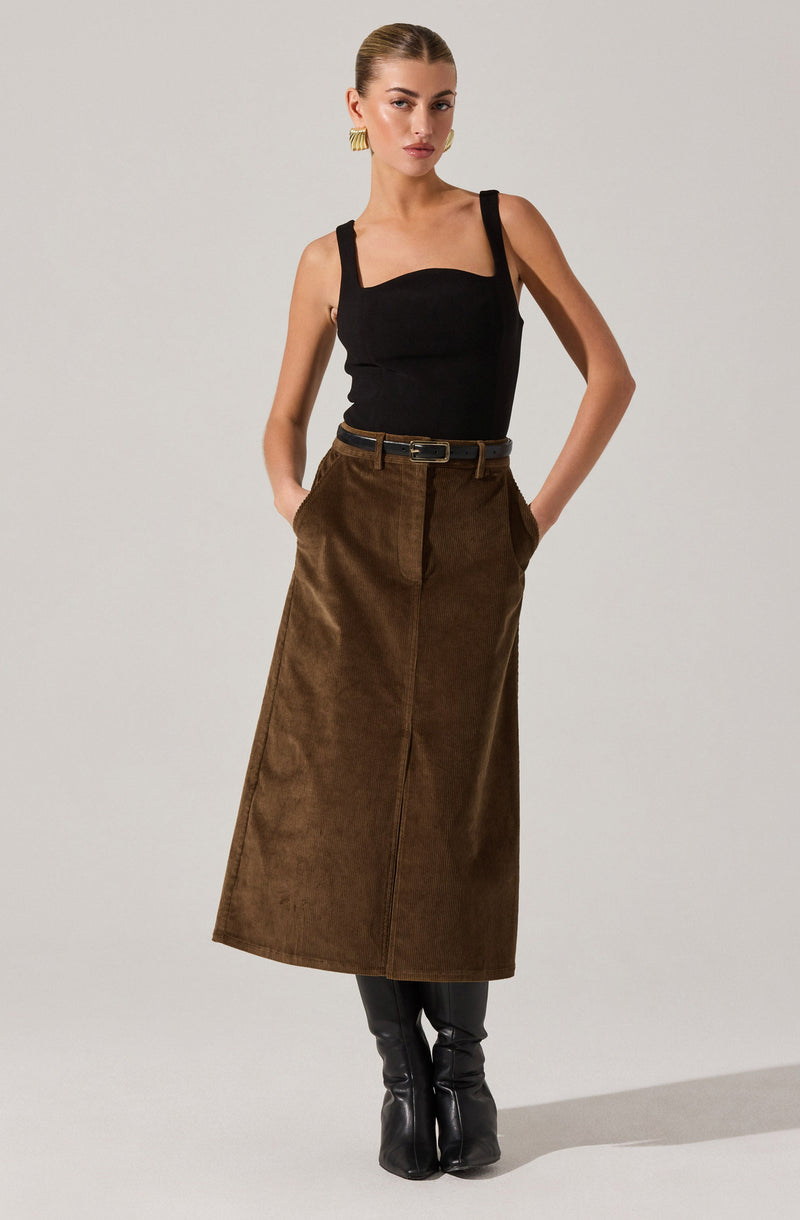 A woman is wearing a black sleeveless top with wide straps and a square neckline, tucked into a high-waisted, brown, midi-length skirt made of a textured fabric. She has accessorized with a belt and large gold earrings, and she is wearing black knee-high boots.