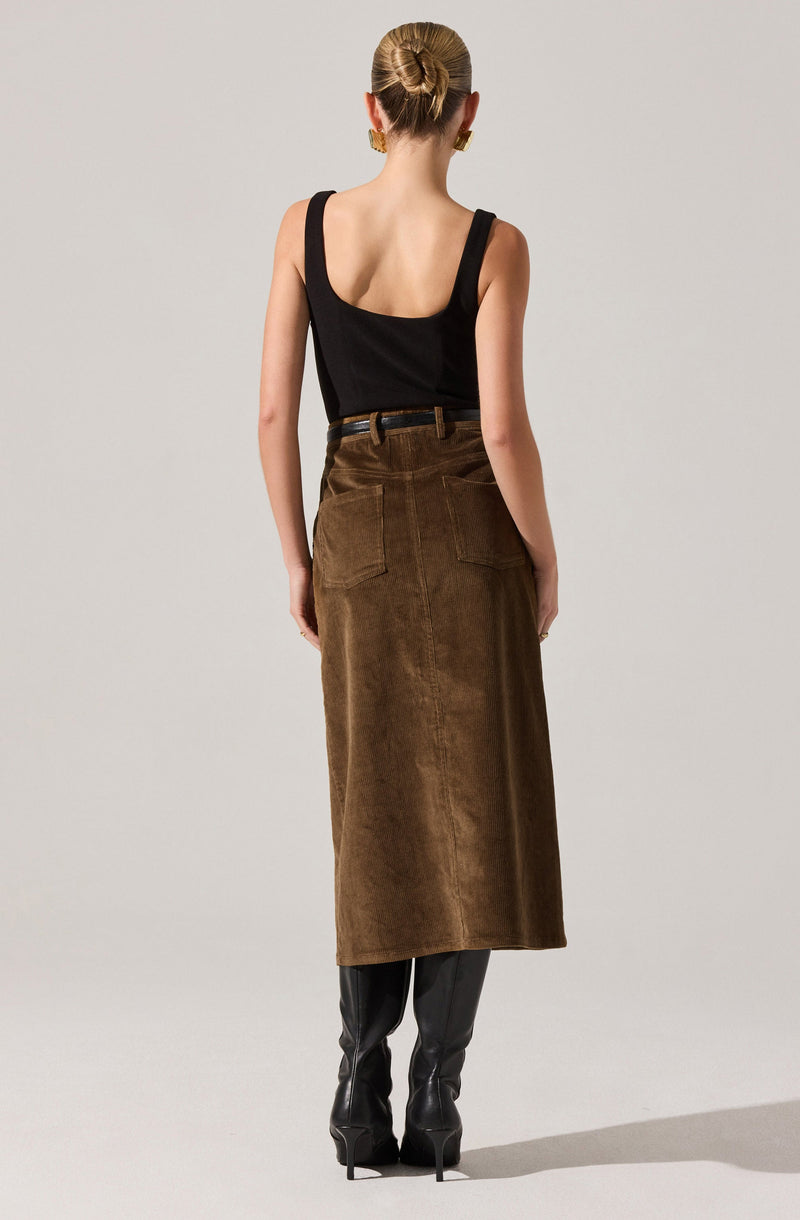 A woman is wearing a black sleeveless top with a wide scoop neckline, tucked into a high-waisted, long brown leather skirt with visible stitching and back patch pockets. She has her hair styled in a low bun and is accessorized with large gold earrings and a thin belt. She is also wearing black high-heeled boots.