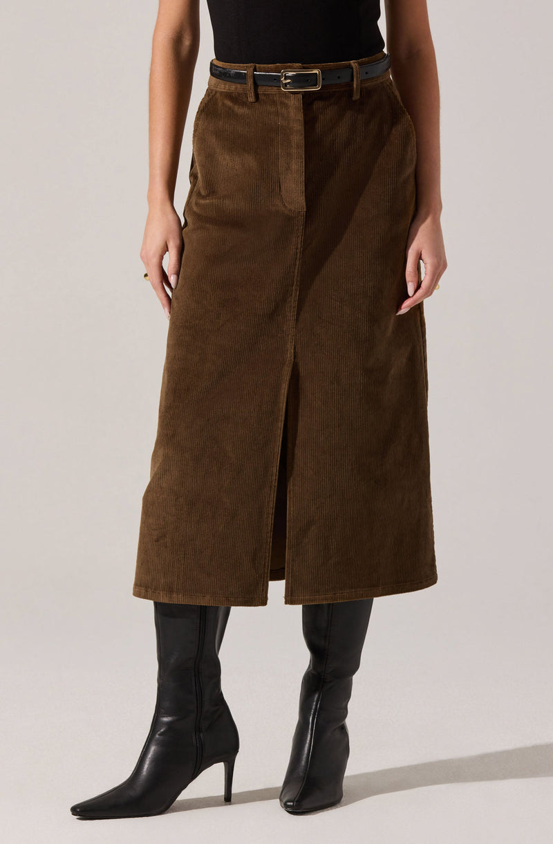A woman is wearing a black sleeveless top tucked into a high-waisted, brown corduroy midi skirt with a front slit. She has a black belt with a gold buckle and is wearing black knee-high boots with pointed toes and high heels.