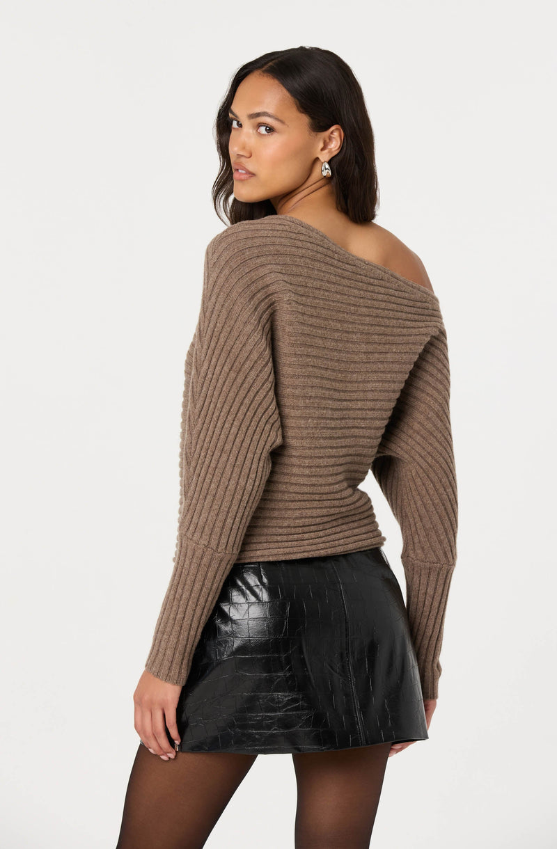 A woman is wearing a loose, off-the-shoulder, ribbed sweater in a tan color. The sweater has wide horizontal ribbing and drops off one shoulder, exposing the collarbone. She pairs it with a black, shiny, crocodile-textured mini skirt and sheer black tights. She also has on silver earrings.