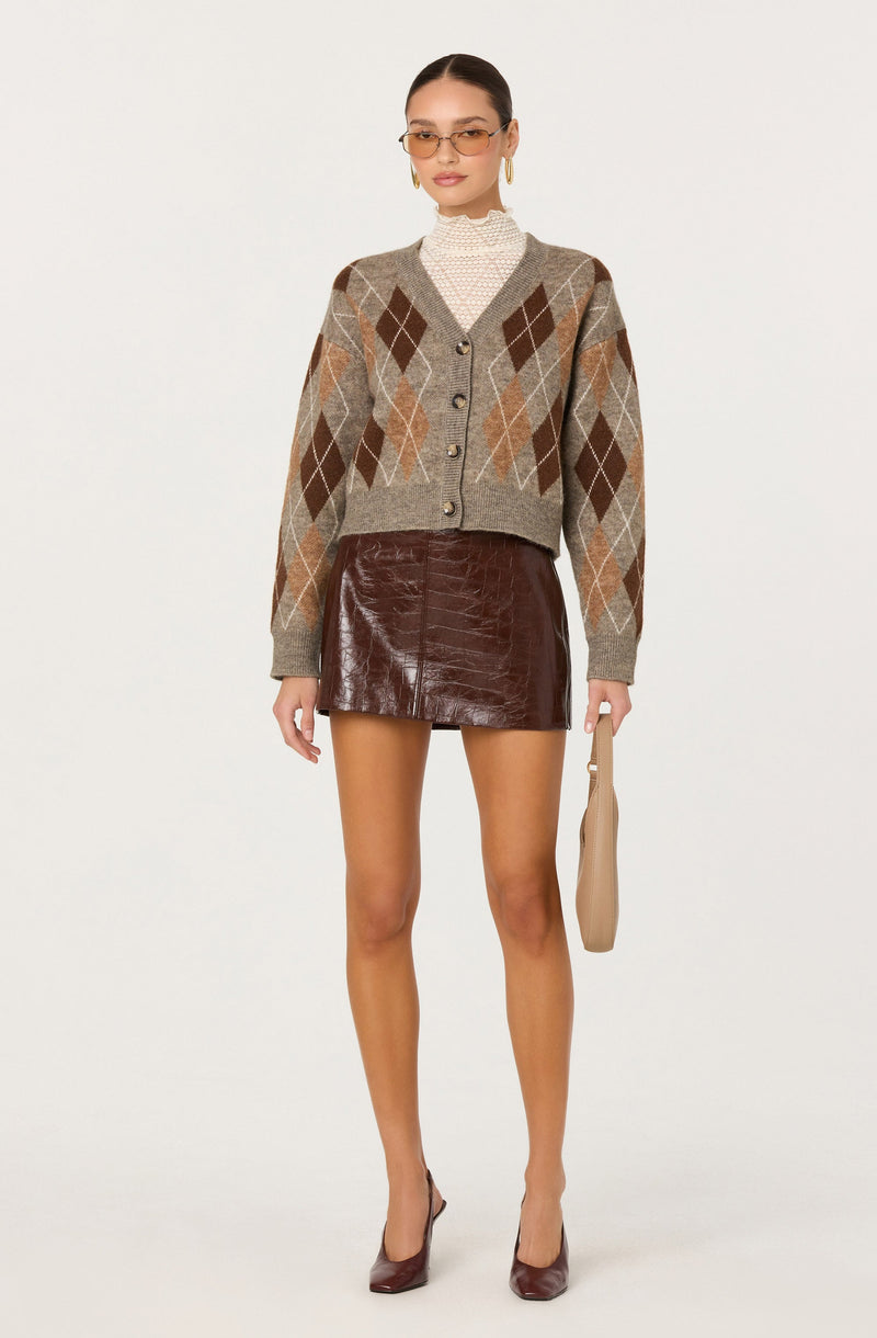 A woman is wearing a beige, brown, and gray argyle patterned cardigan with large buttons. Underneath, she has on a high-neck, white, textured top. She pairs the outfit with a shiny, dark brown leather mini skirt. She accessorizes with gold hoop earrings, sunglasses, and holds a beige clutch bag. She completes the look with matching burgundy high heels.