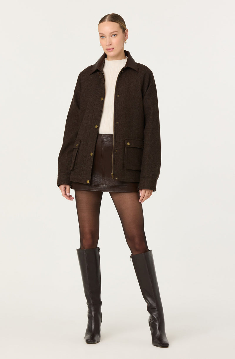 A woman is wearing a dark brown, oversized coat with a collar and button closures. Underneath, she has on a white, knitted sweater. She is also wearing a dark brown leather mini skirt and sheer black tights. Completing the outfit, she has on tall, black leather knee-high boots.