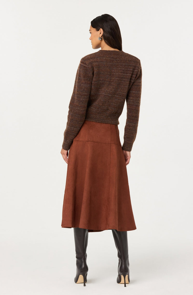 A woman is wearing a long brown knitted sweater with multicolored horizontal stripes and a high neckline, paired with a high-waisted, rust-colored, suede A-line midi skirt. She is also wearing tall, black leather heeled boots.
