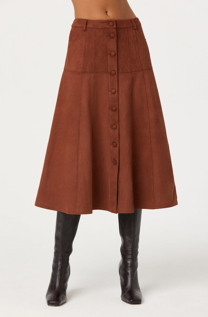 A woman is wearing a high-waisted, brown, midi-length skirt with a front button closure. The skirt features visible vertical and horizontal seam details, giving it a structured appearance. She is also wearing tall, black leather boots that reach just below the knees.