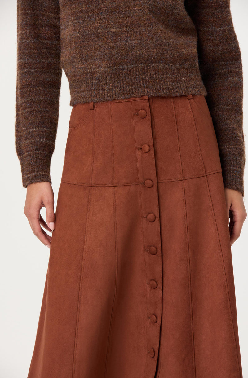 A person is wearing a brown, long-sleeved, textured sweater and a high-waisted, rust-colored leather skirt with vertical and horizontal seam details. The skirt has a row of fabric-covered buttons running down the front center.