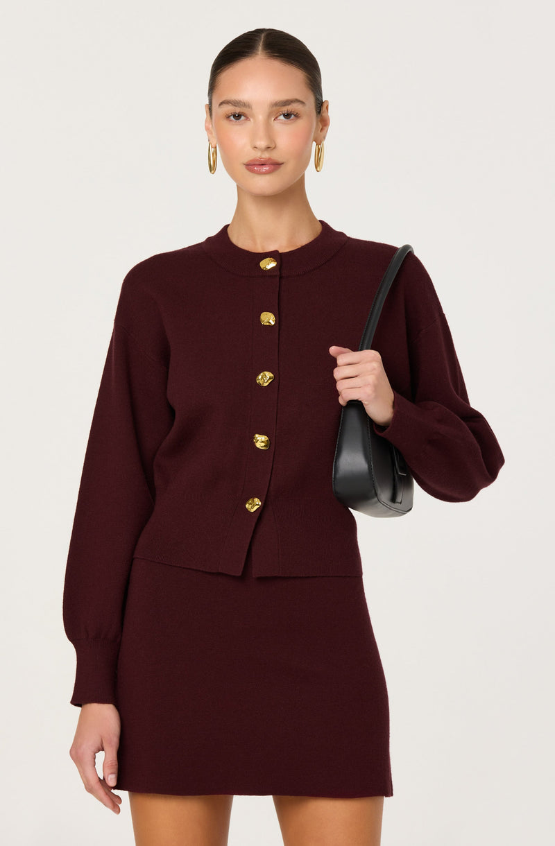 A woman is wearing a burgundy knit outfit consisting of a long-sleeved, button-up cardigan with gold buttons and a matching fitted skirt. She has gold hoop earrings and is carrying a black handbag over her shoulder.