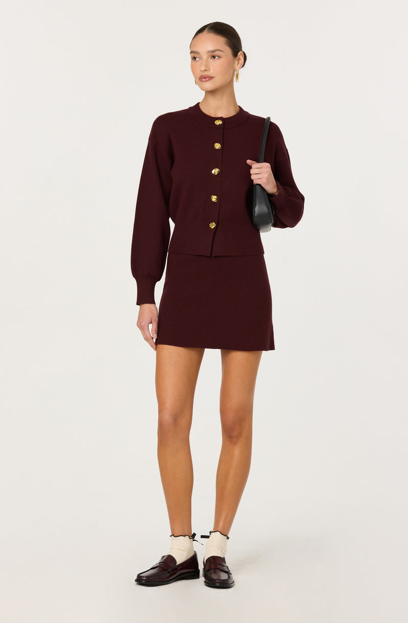 A woman is wearing a matching burgundy outfit consisting of a long-sleeve, button-up cardigan with gold buttons and a matching fitted mini skirt. She has her hair pulled back and is accessorizing with gold hoop earrings and carrying a black shoulder bag. She is also wearing white socks and dark brown loafers.