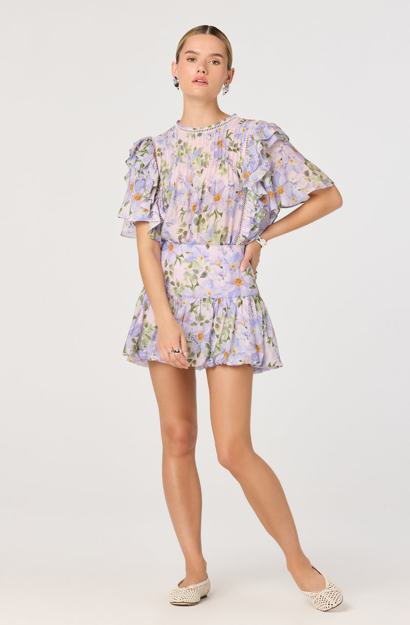 The outfit consists of a matching two-piece set with a floral pattern in pastel purple, green, and yellow hues. The top features ruffled short sleeves and a round neckline with decorative trim, while the high-waisted shorts have a ruffled hem that creates a playful, layered look. The fabric appears lightweight and textured, suitable for warm weather. The model is also wearing light-colored, woven flat shoes.