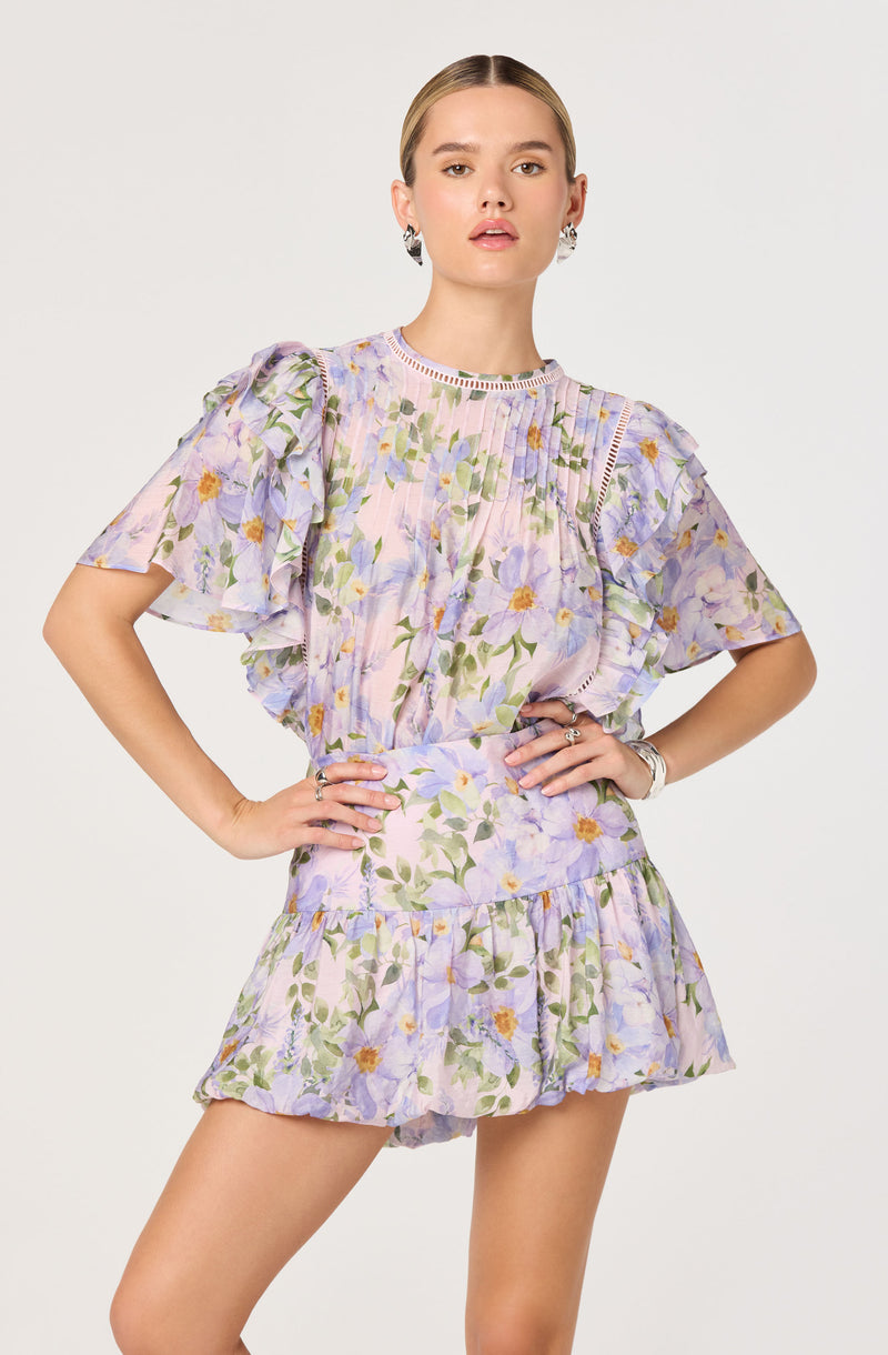 A woman is wearing a matching floral-printed top and shorts set. The top has a pastel purple base with a watercolor-style lavender, green, and yellow floral pattern. It features puffed short sleeves with ruffle detailing and a round neckline with intricate trim accents. The matching shorts have the same purple floral pattern, a ruffled hem, and a high waist.