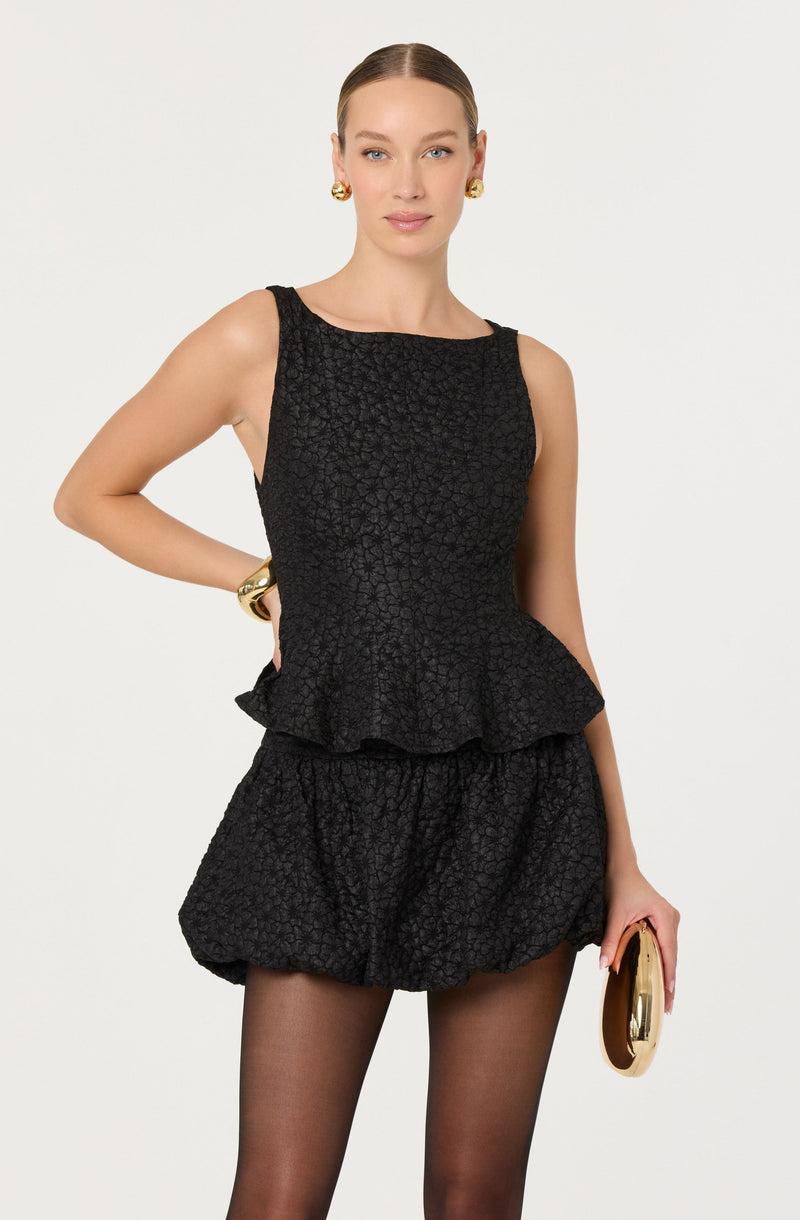 A woman is wearing a sleeveless black textured peplum top and matching textured shorts. She has gold hoop earrings, a gold bracelet, and is holding a gold clutch in her right hand. She has her hair parted in the middle and styled straight.
