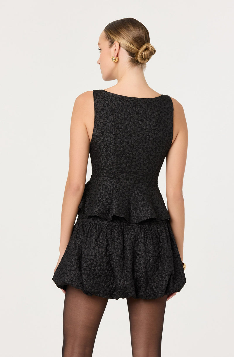 A woman is wearing a sleeveless black top with a textured, floral pattern and a peplum hem. She pairs it with matching black textured shorts that are loose-fitting and have a gathered, balloon style. She is also wearing sheer black tights, gold earrings, and a gold bracelet.