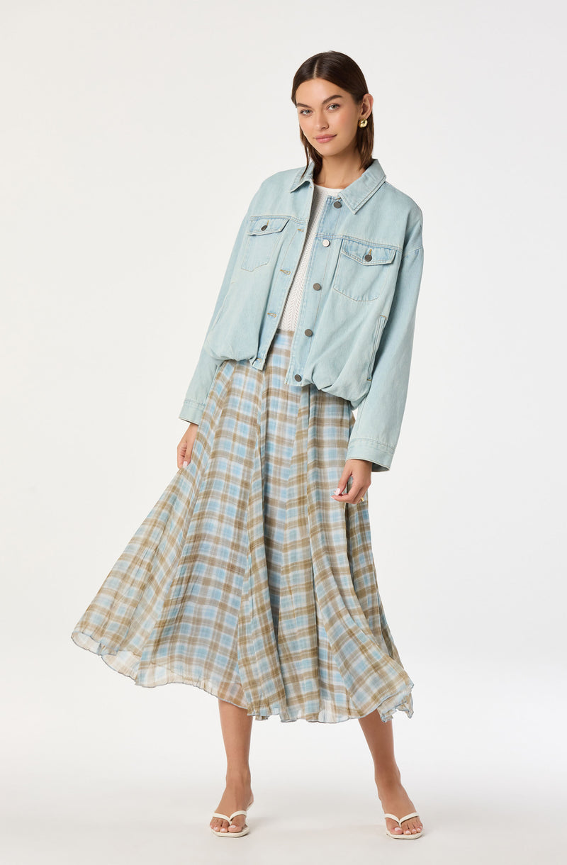 Woman wears a light blue denim jacket, beige and blue plaid skirt, and white sandals.