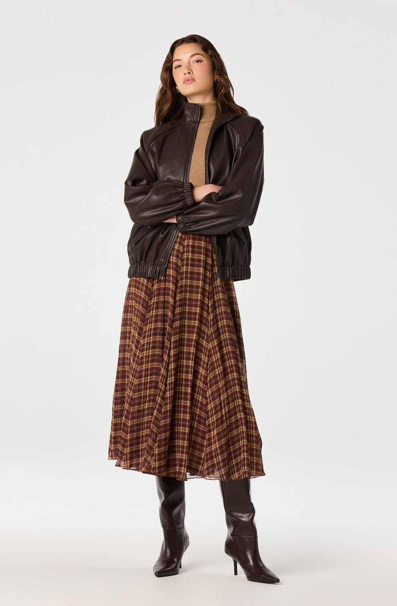 Woman wearing a dark brown leather jacket, a tan top, a plaid brown skirt, and tall black boots.