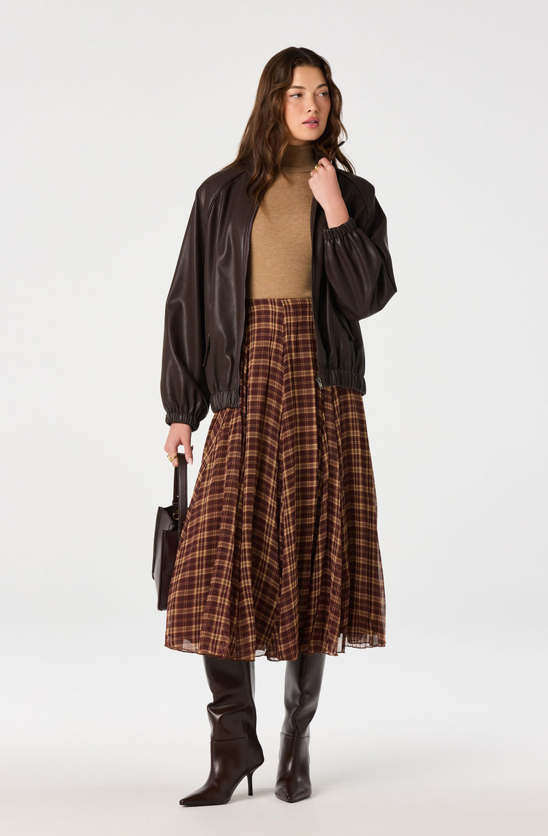 Woman wearing a dark brown leather bomber jacket, a tan sweater, a plaid brown and red midi skirt, black high-heeled boots, and holding a black handbag.