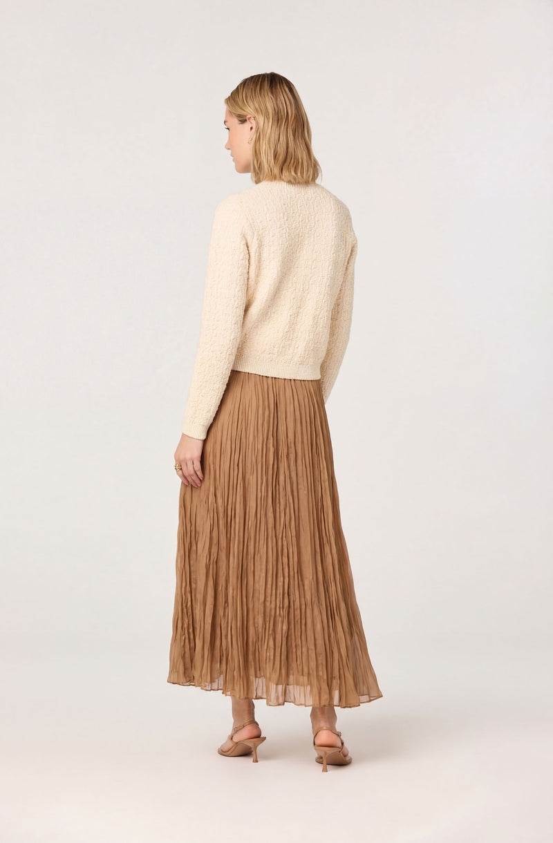 Woman wearing a cream textured sweater and a long, pleated, light brown skirt.