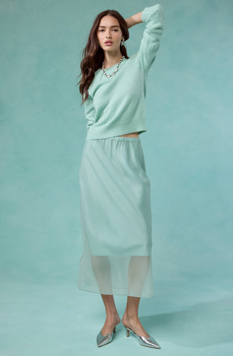 A woman is wearing a light mint green, long-sleeved knit sweater with a relaxed fit and a ribbed hem, paired with a matching mint green satin midi skirt that is slightly shiny and has an elastic waistband. She is also wearing silver metallic pointed-toe high heels with a small heel, and a pearl necklace.