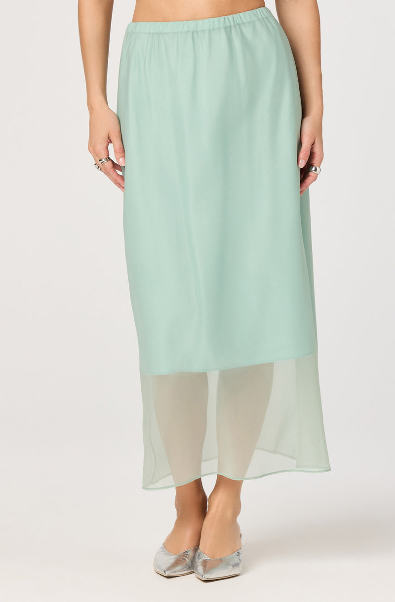 A person is wearing a long, light mint green skirt with an elastic waistband. The skirt has a sheer, translucent overlay that reaches to the ankles, creating a layered look. The person is also wearing metallic silver pointed-toe shoes.