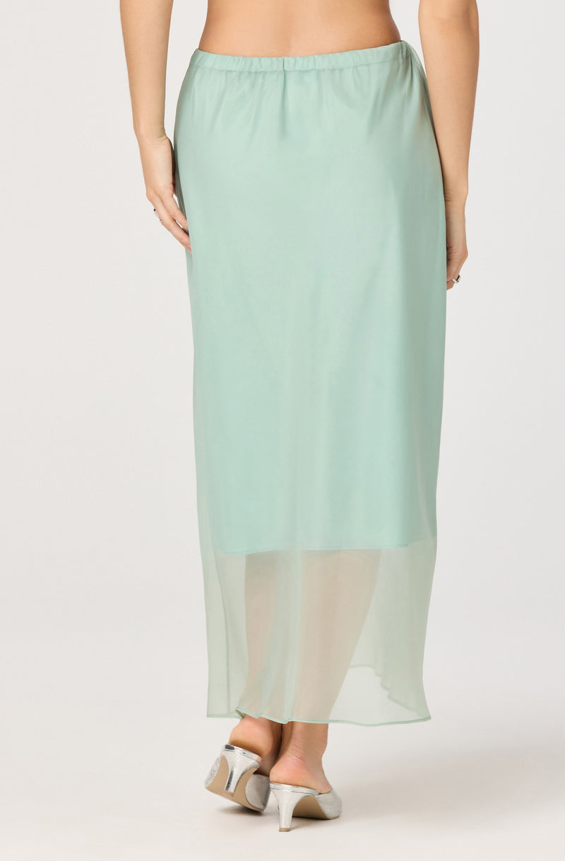 A person is wearing a long, light green, semi-transparent skirt with an elastic waistband. The skirt has a flowing, lightweight fabric that reaches the ankles. They are also wearing white, open-toe heels with a block heel.