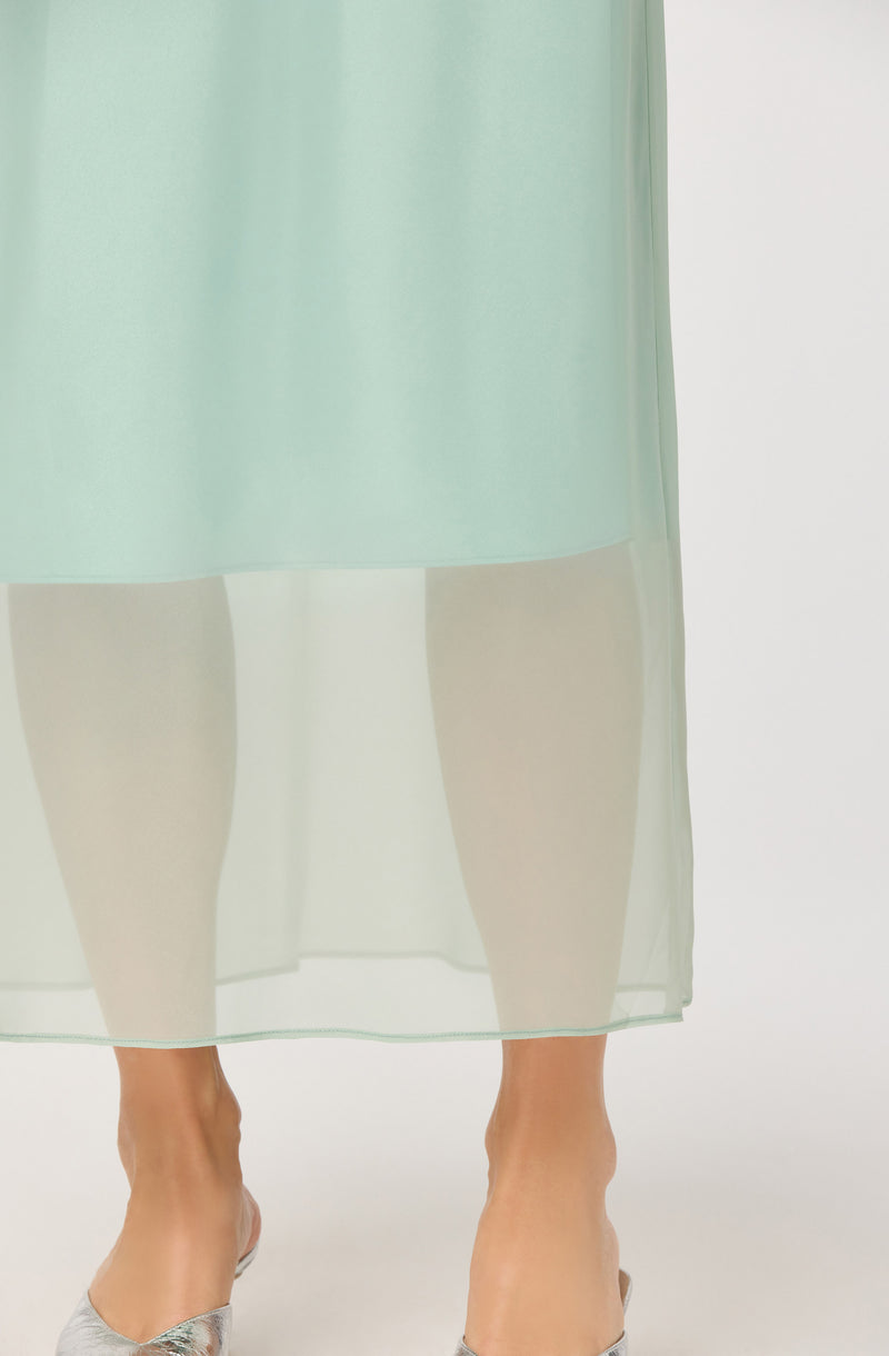 The image shows a woman wearing a light mint green dress made of a sheer, flowy fabric that extends below the knee. She is also wearing metallic silver pointed-toe heels.