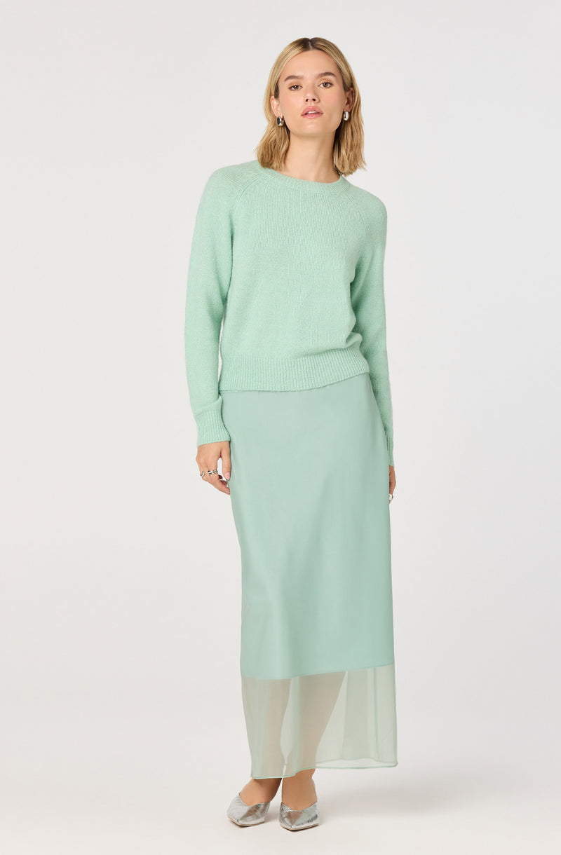 A woman is wearing a light mint green crewneck sweater with long sleeves and ribbed cuffs, paired with a matching long, flowy skirt that has a sheer overlay at the bottom. She is also wearing metallic silver pointed-toe shoes.