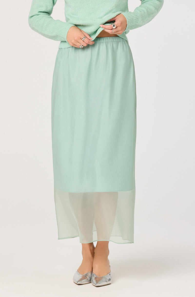 A person is wearing a light green sweater with long sleeves and a high neckline. They are also wearing a long, light green satin skirt with a sheer, semi-transparent layer at the bottom that reaches their ankles. They are wearing silver pointed-toe flats with a shiny finish. The person has multiple rings on both hands.