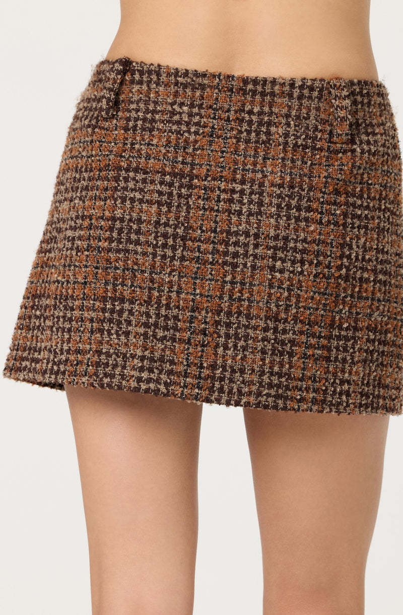 A woman is wearing a short, high-waisted skirt made of a textured, woolen tweed fabric. The skirt features a classic houndstooth pattern in shades of brown, black, and tan, with small checks throughout. It has a tailored fit with belt loops at the waistband.