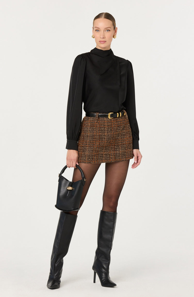 A woman is wearing a black long-sleeve top with a high neckline, slightly puffed shoulders, and fitted cuffs. She has on a brown and black plaid mini skirt with a textured pattern, paired with a black belt featuring a gold buckle. She is also wearing sheer black tights and tall black leather boots with high heels. She holds a small black handbag with a gold detail in her right hand.