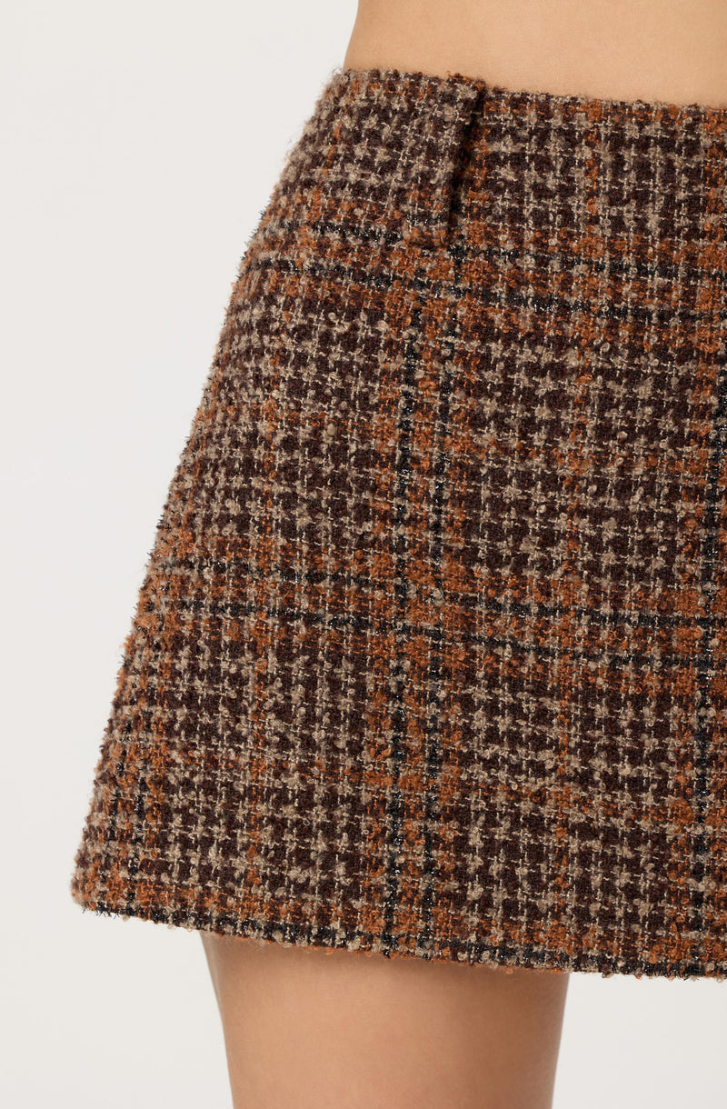 A close-up of a skirt made from a textured, brown and orange plaid tweed fabric. The skirt has a straight cut and features a waistband with a subtle slit at the top. The fabric appears warm and suitable for fall or winter wear.