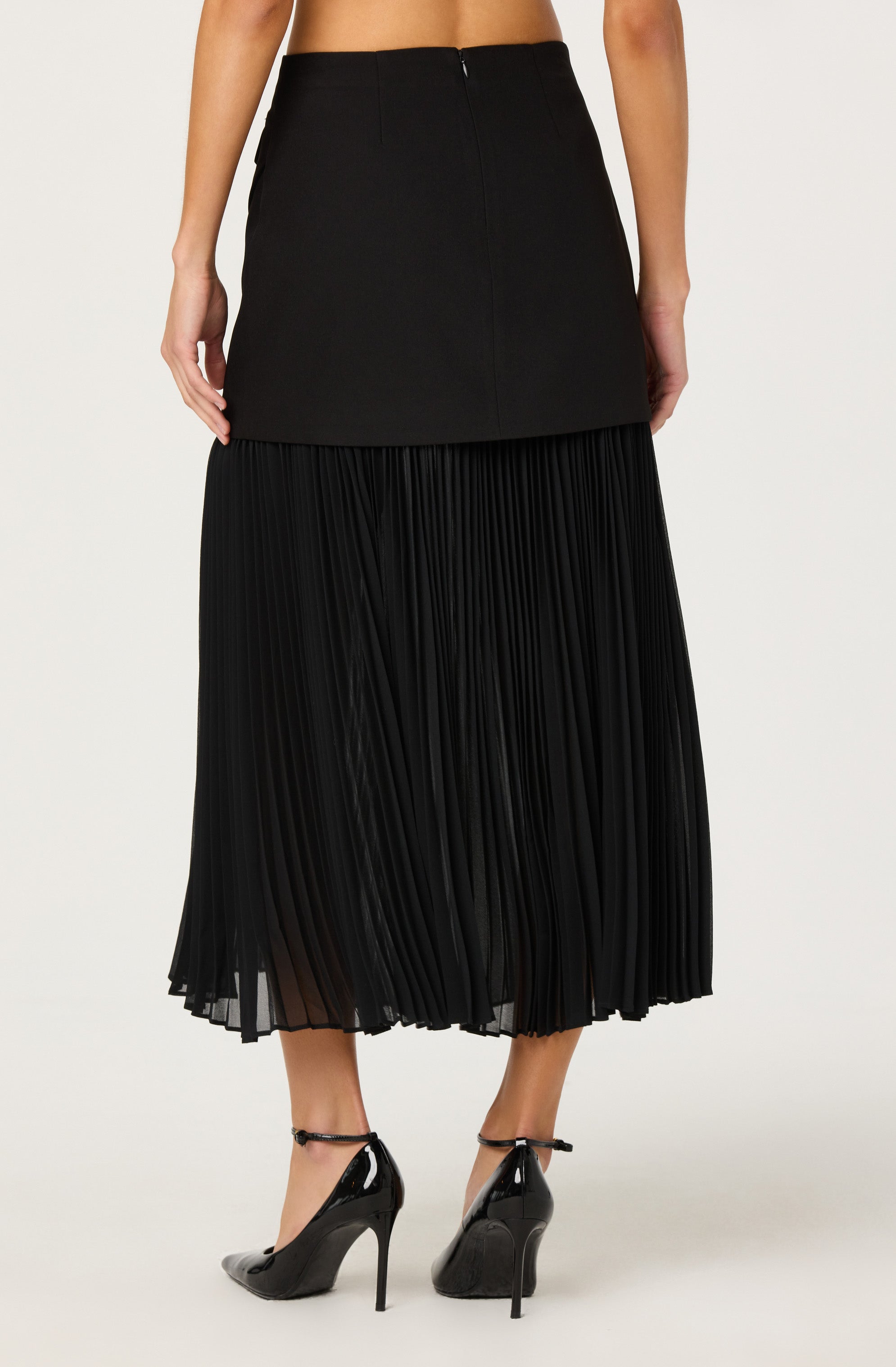 A woman is wearing a black high-waisted skirt with a straight, fitted top section and a pleated, flowing lower section that extends to her mid-calf. She is also wearing black patent leather high-heeled shoes with ankle straps.