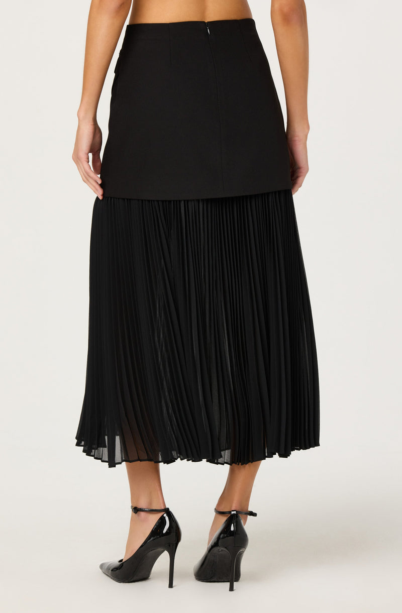 A woman is wearing a black high-waisted skirt with a straight, fitted top section and a pleated, flowing lower section that extends to her mid-calf. She is also wearing black patent leather high-heeled shoes with ankle straps.