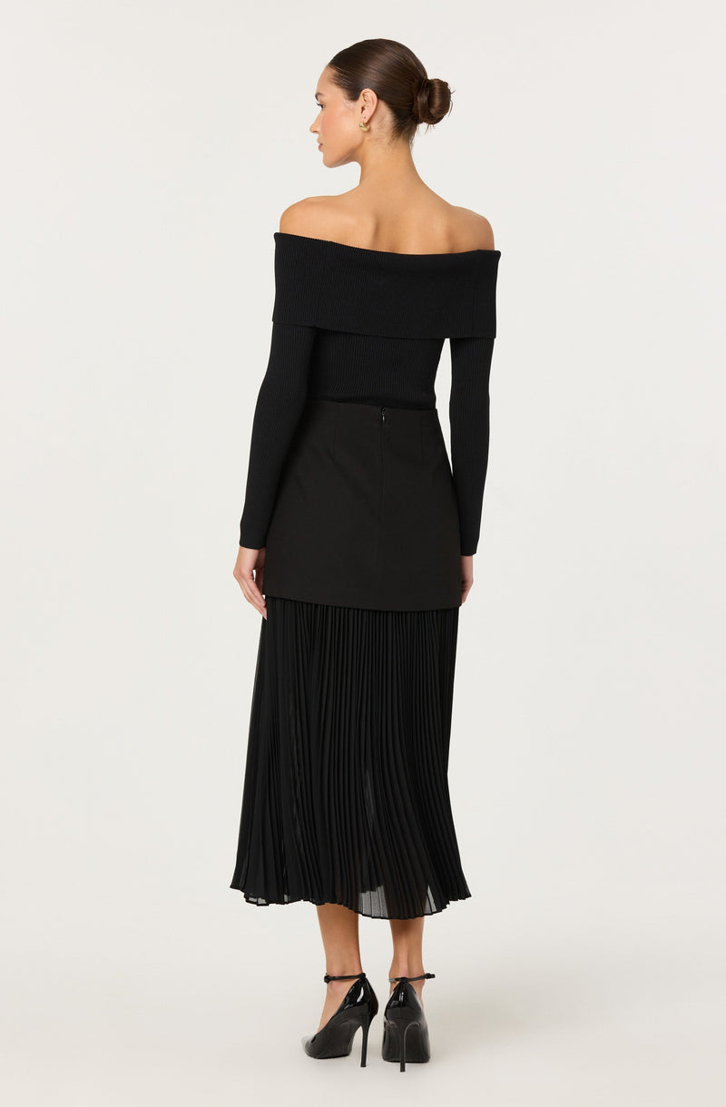 A woman is wearing an off-the-shoulder black sweater with long fitted sleeves, paired with a high-waisted black skirt that has a pleated, flowy design reaching mid-calf length. She is also wearing black high-heeled shoes with ankle straps. Her hair is styled in a neat bun.