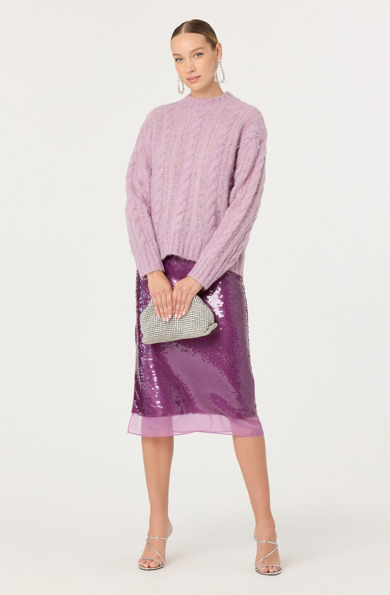 A woman is wearing a soft, oversized, pastel pink cable-knit sweater with a high neckline and long sleeves. She is also dressed in a shiny, purple sequined midi skirt that reaches just below her knees, with a sheer pink hem at the bottom. She accessorizes with large dangling earrings, a silver beaded clutch purse, and strappy silver high-heeled sandals.