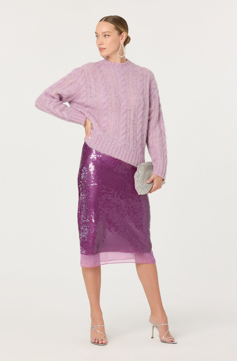 A woman is wearing a pastel pink, chunky cable-knit sweater with a high neckline and long sleeves that extend past her wrists. She pairs it with a shiny, sequined purple midi skirt that has a sheer pink hem at the bottom. She accessorizes with large pearl earrings, holding a small, silver, beaded clutch, and wears strappy high-heeled sandals.