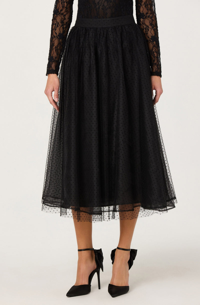A woman is wearing a black lace long-sleeve top with a floral pattern and a high neckline. She has a black, high-waisted midi skirt made of sheer tulle fabric with small polka dots, layered over a lining. She is also wearing black high-heeled shoes with ankle straps tied into bows.